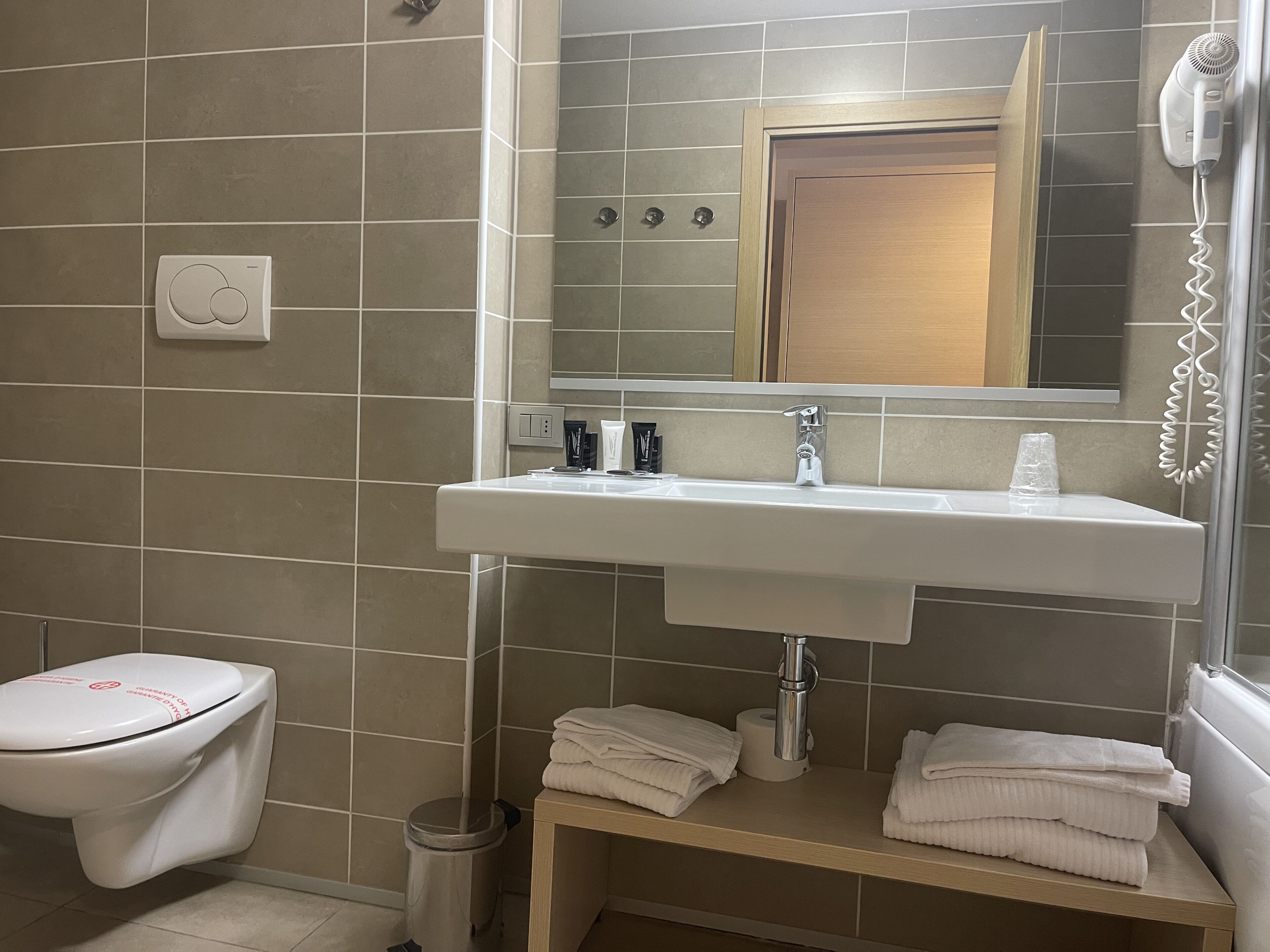 A bathroom with a toilet, sink, and a mirror above it. There is a towel rack with towels and a trash can.