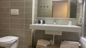 A bathroom with a toilet, sink, and a mirror above it. There is a towel rack with towels and a trash can.