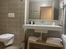 A bathroom with a toilet, sink, and a mirror above it. There is a towel rack with towels and a trash can.