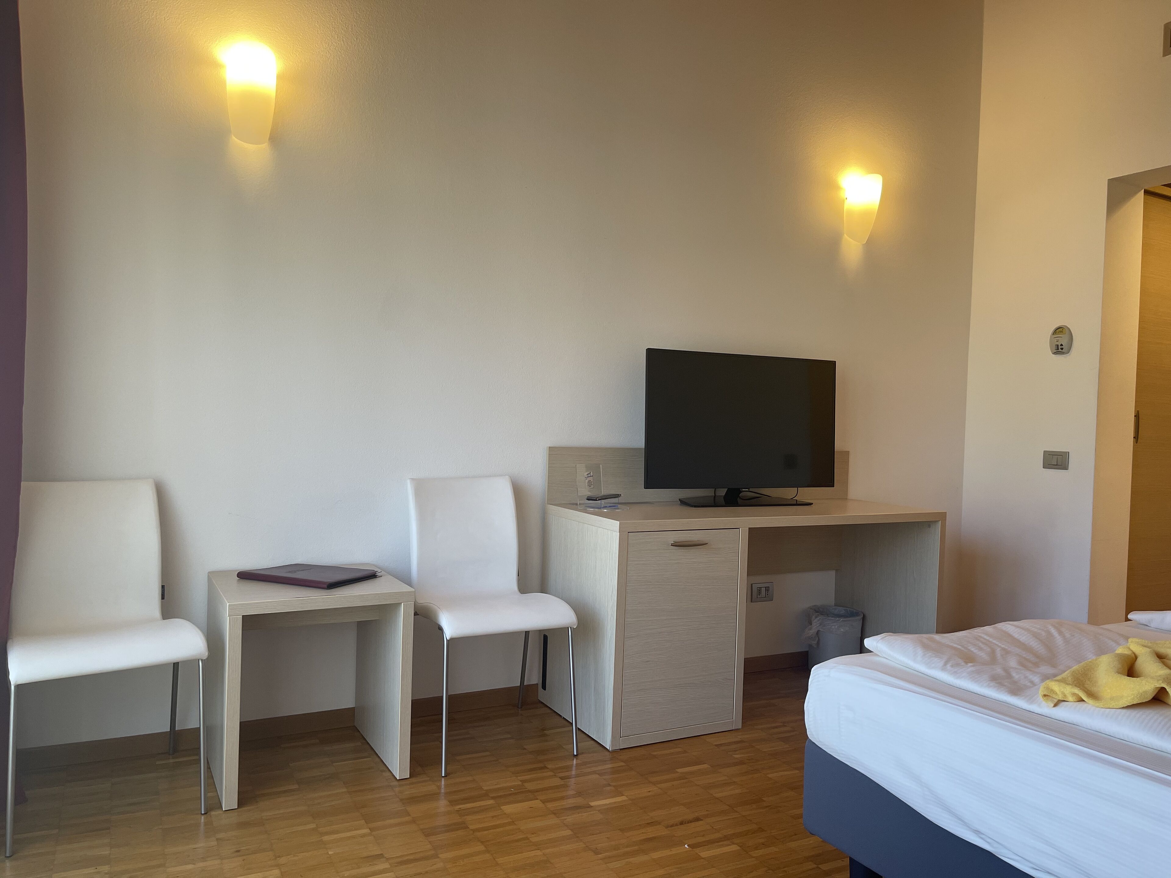 A hotel room with a bed, a desk with a computer, two white chairs, and two wall-mounted lights.