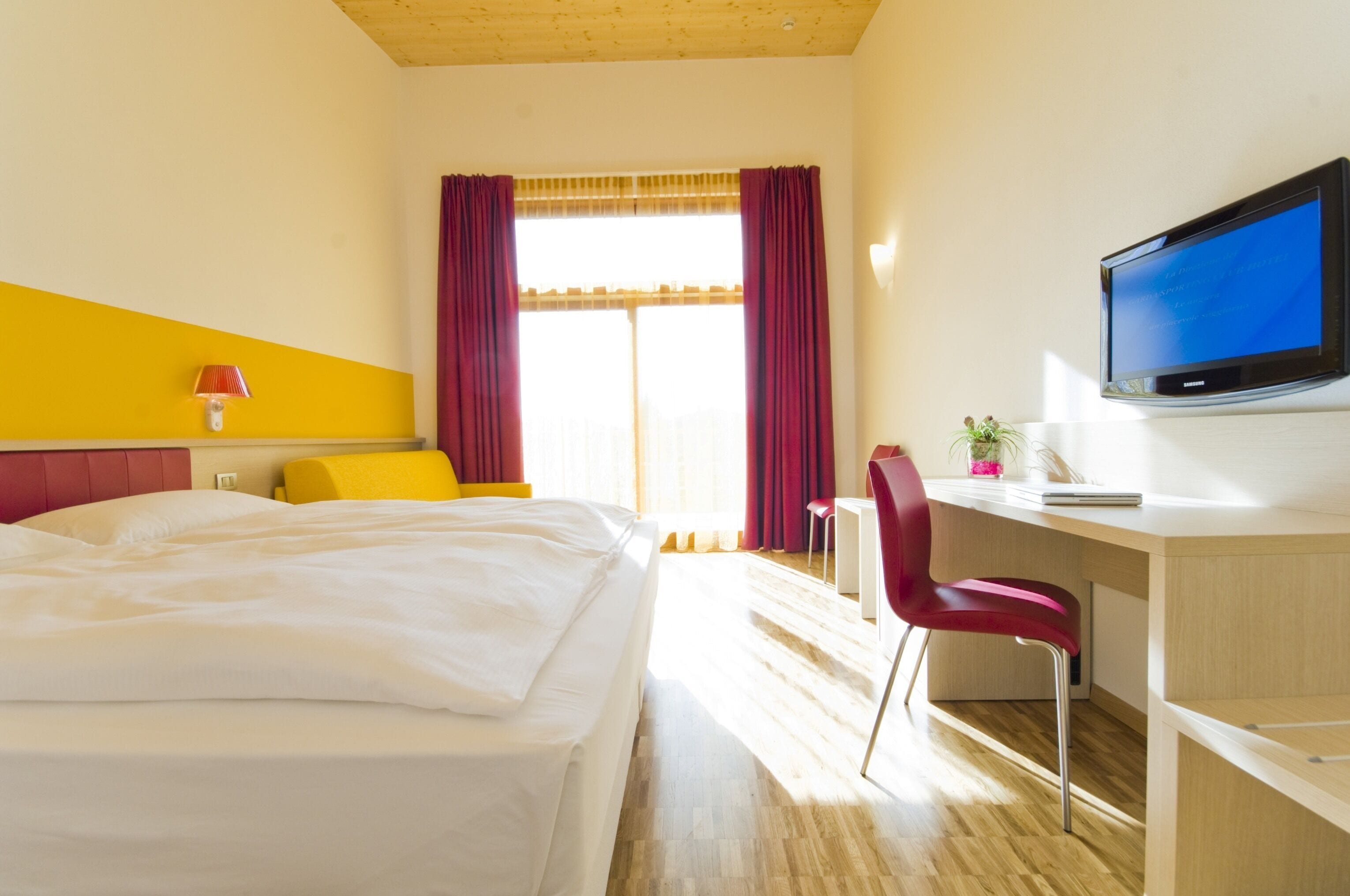 Habitación de hotel con cama, escritorio, silla, televisor y ventana con cortinas.
