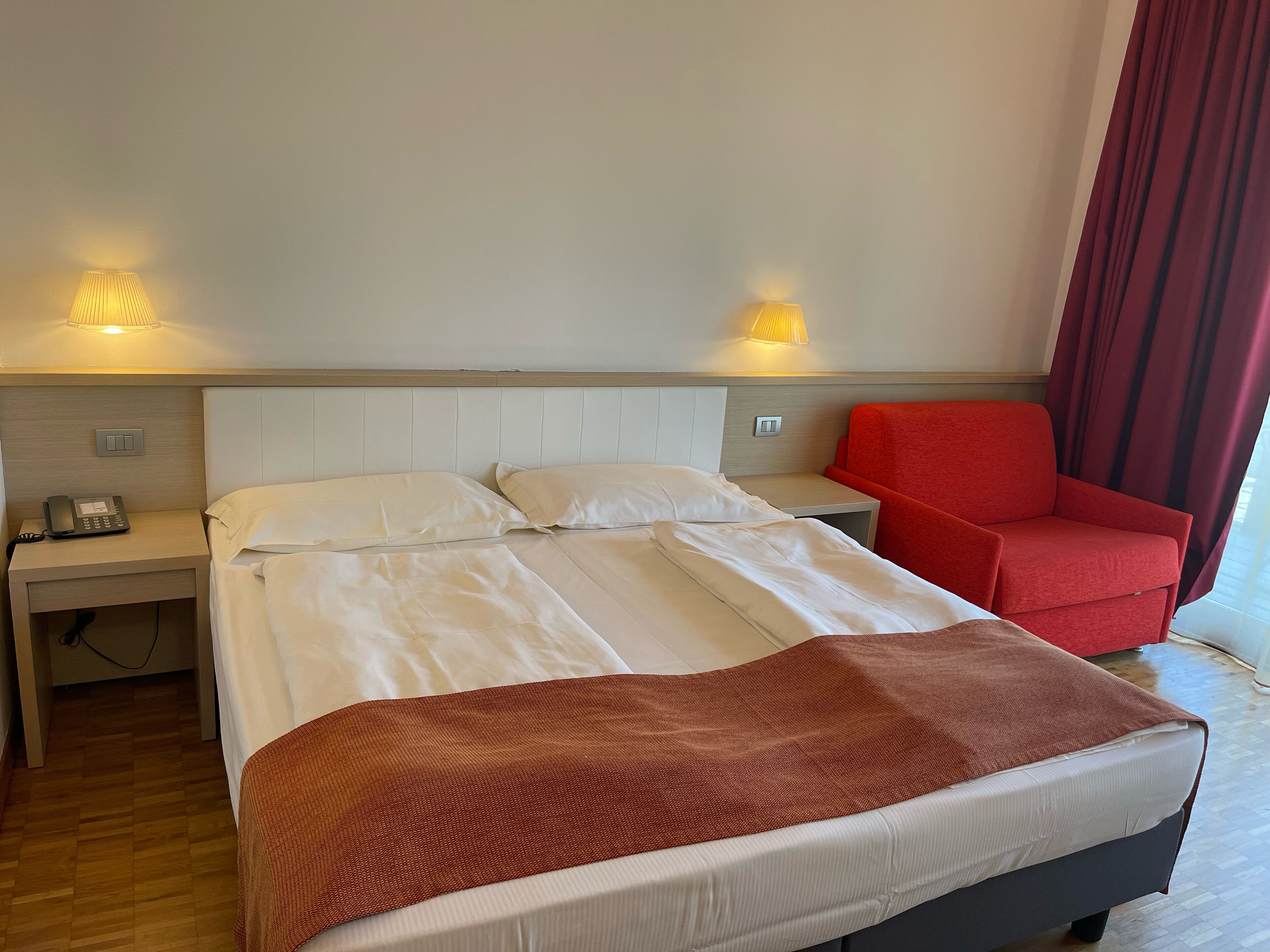 Habitación de hotel con cama, una silla roja, lámparas de noche y un teléfono en la mesita.