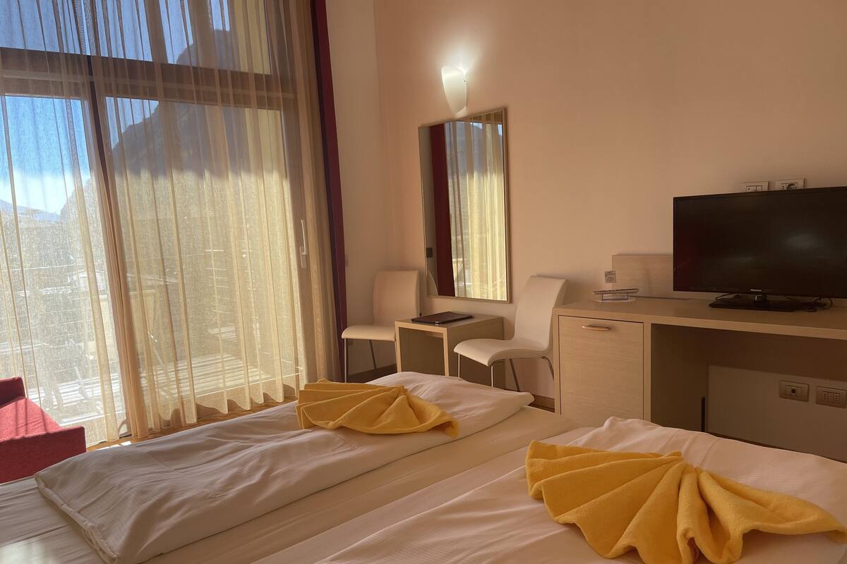 A hotel room with two beds, a desk, a chair, a television, and a large window with sheer curtains.