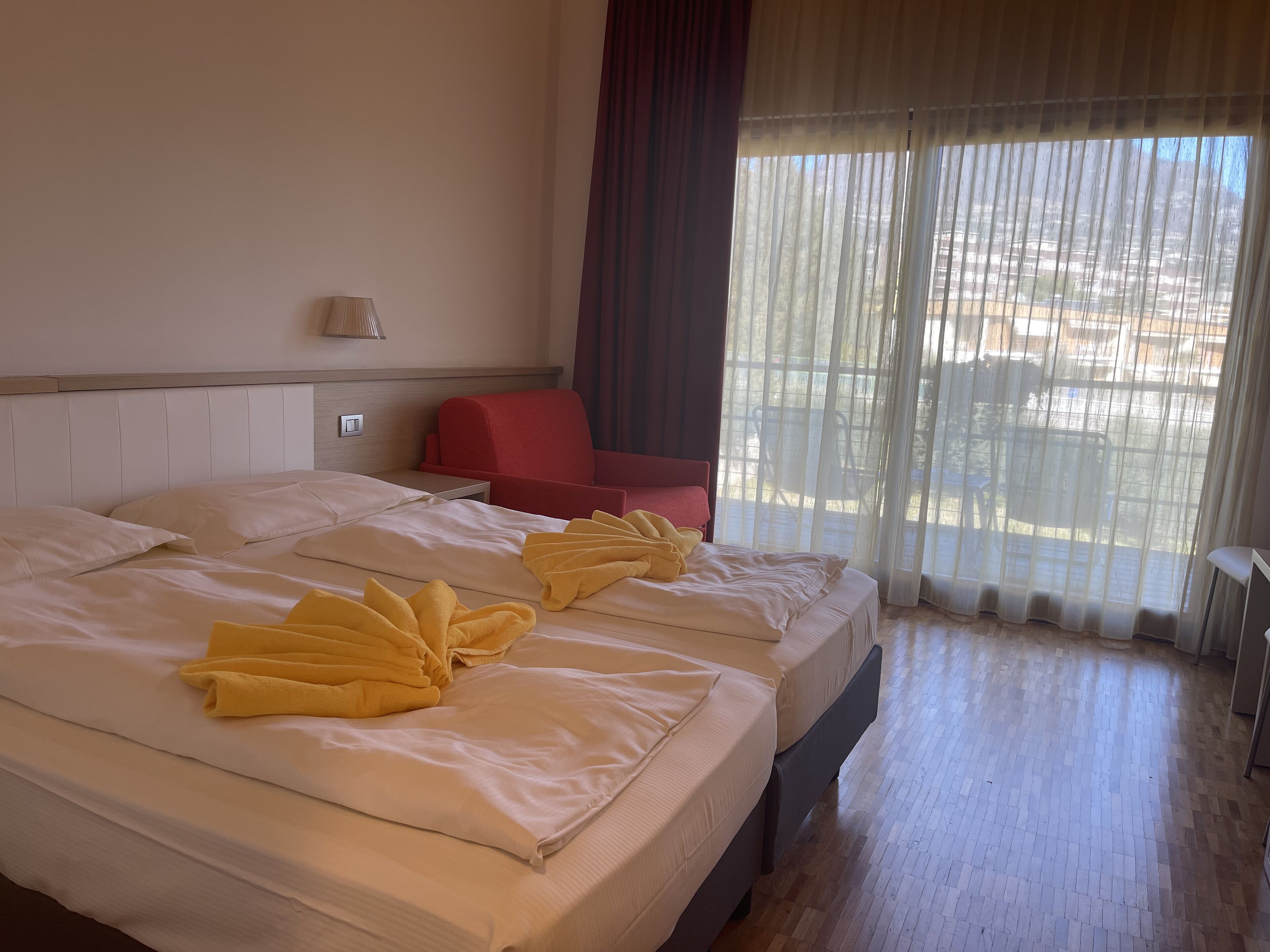 A hotel room with two beds, a red chair, a lamp, and a view through sheer curtains.