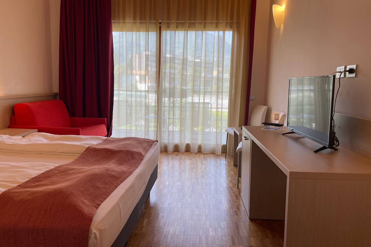 A hotel room with a bed, a desk with a TV, a chair, and a window with curtains.