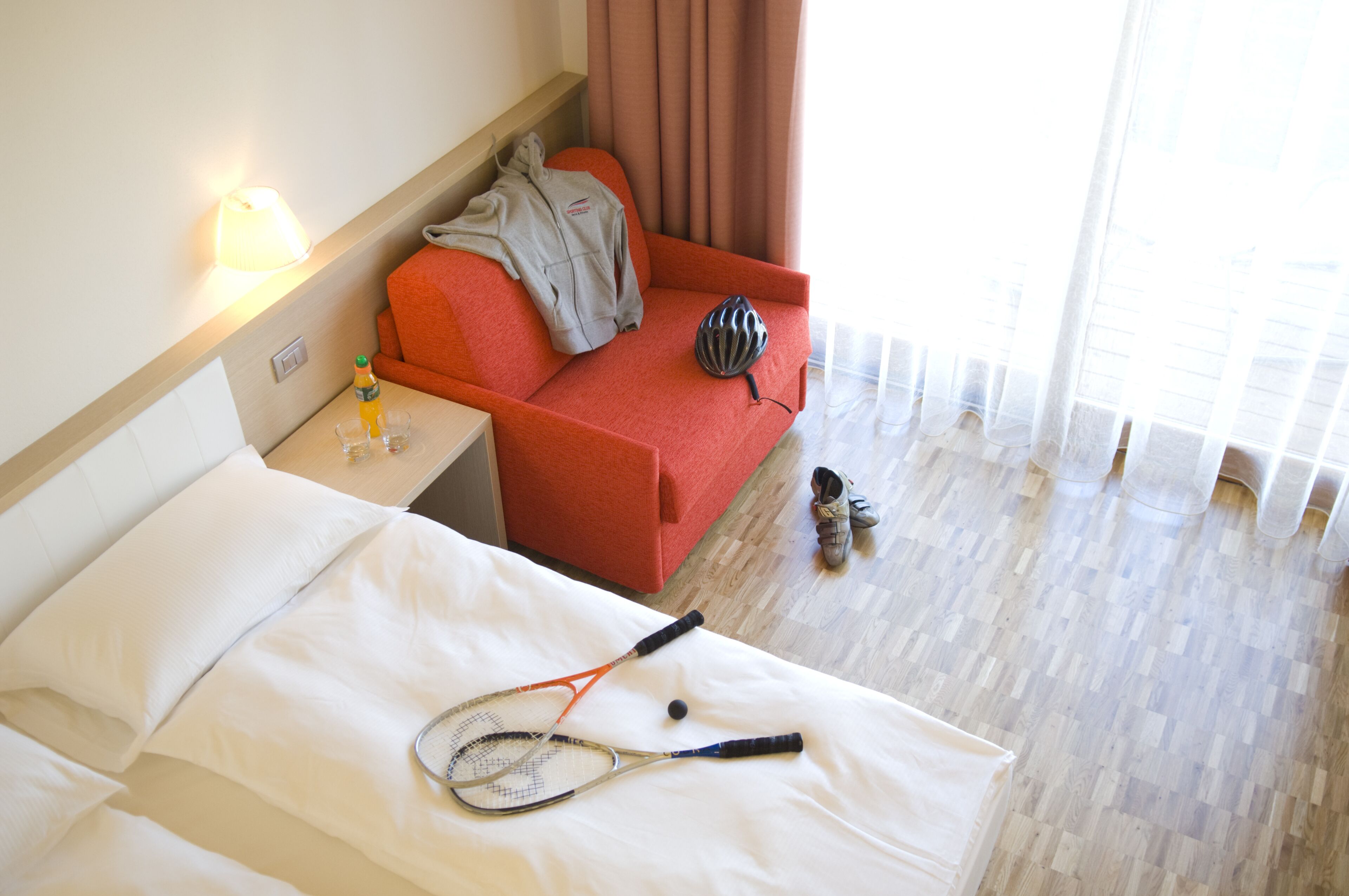 Una habitación de hotel con una cama, un sofá rojo, una raqueta de tenis y un casco en el suelo.