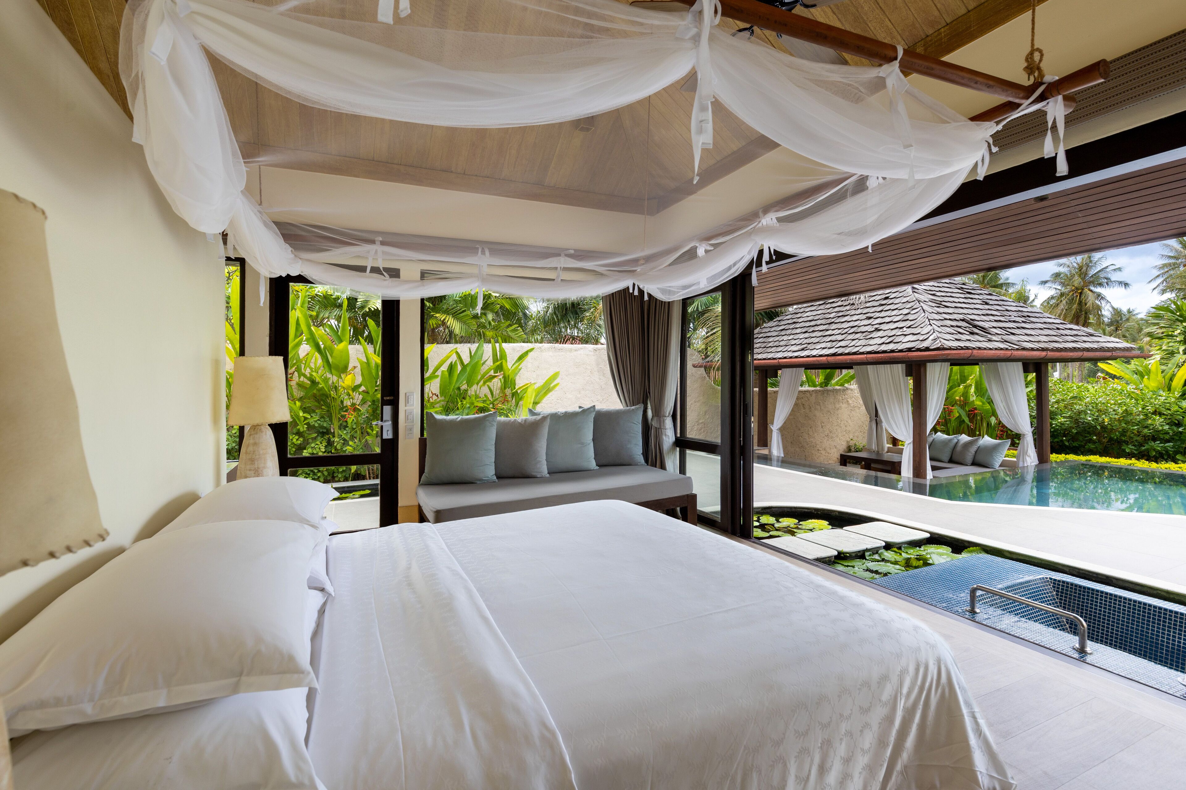 A bedroom with a large bed, a bench with cushions, and a view of an infinity pool.