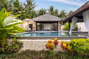 Un área junto a la piscina con una zona de estar techada, una piscina y exuberante follaje tropical.