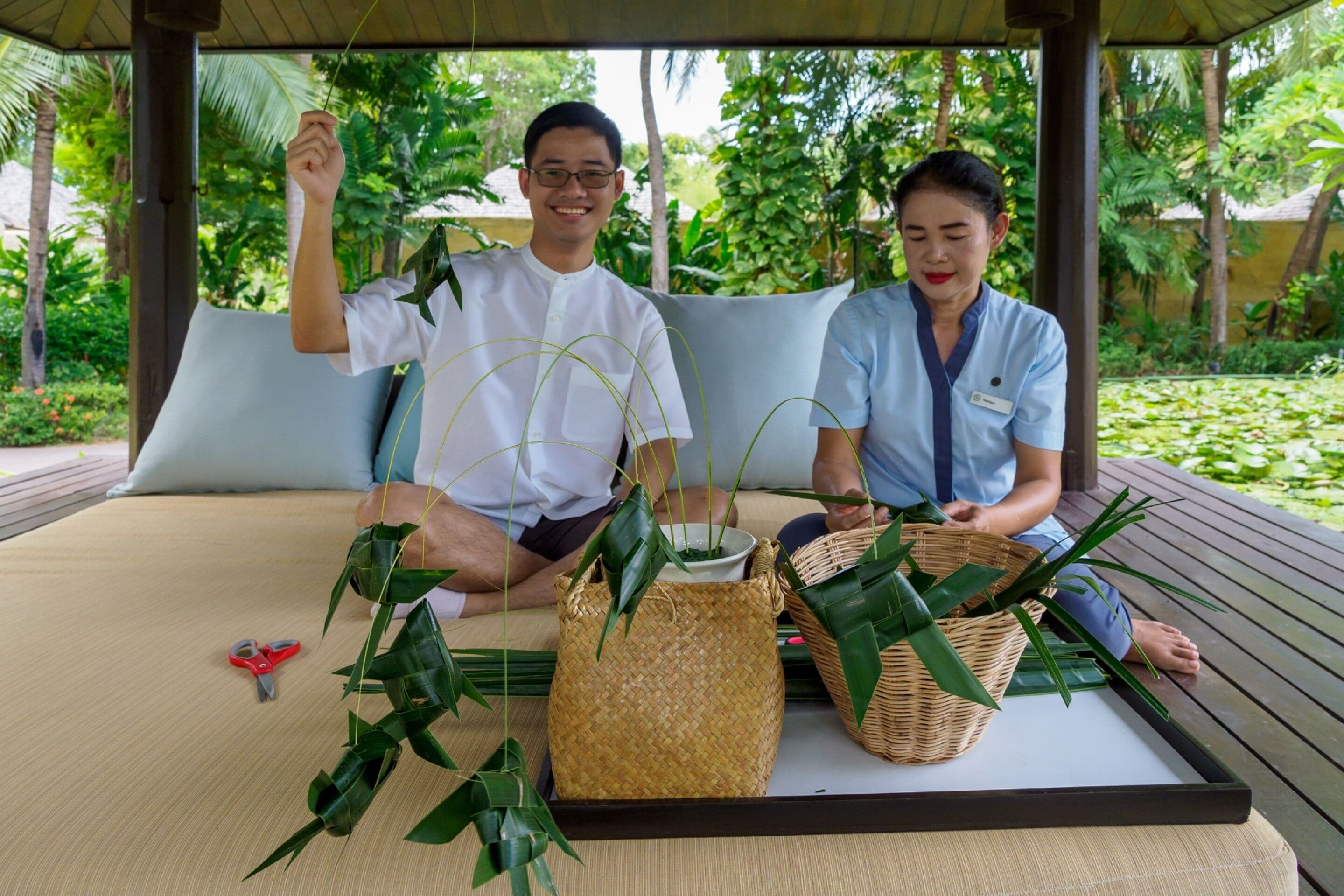 A couple sitting on a bed with woven baskets and green leaves.