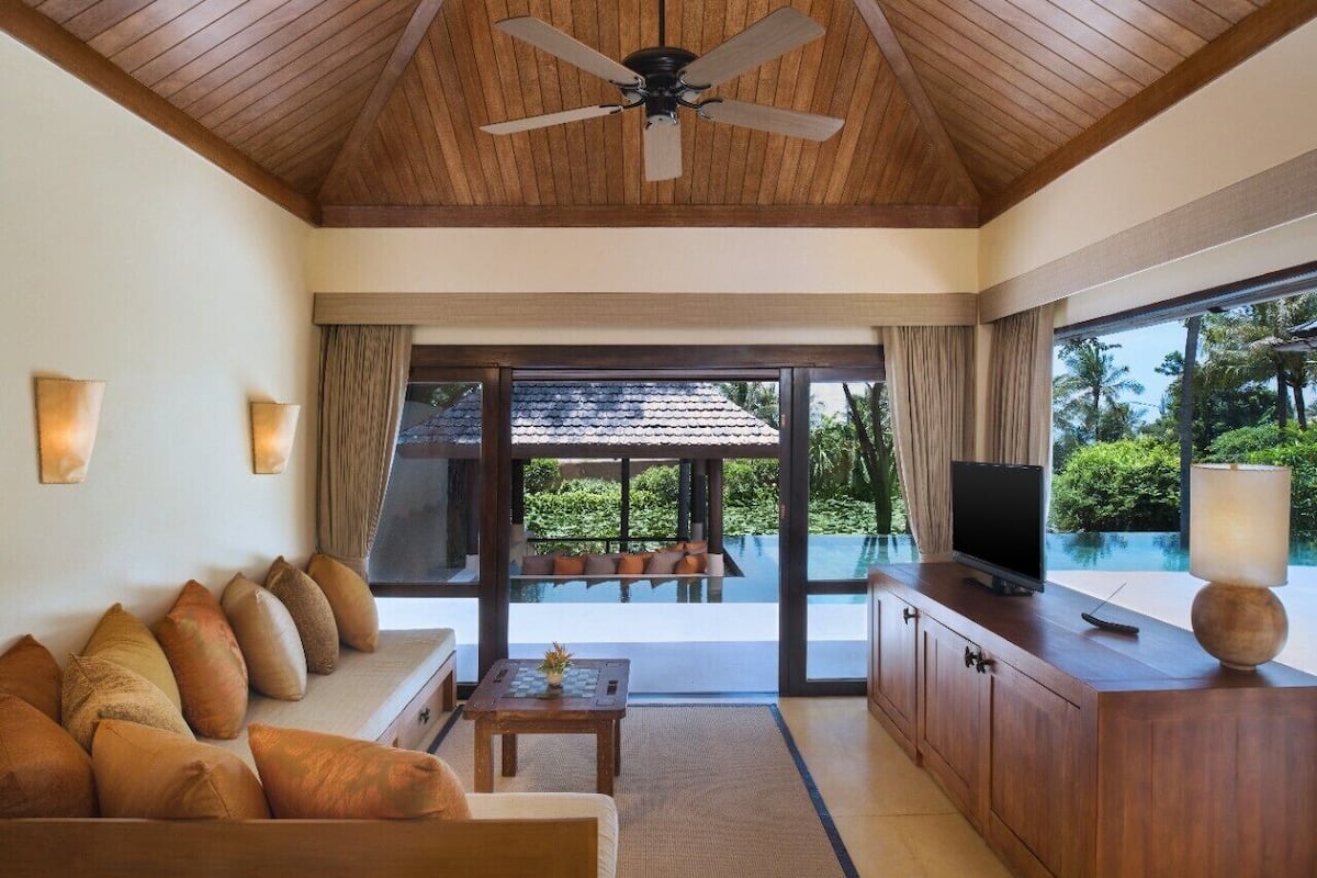 A modern living room with a wooden ceiling, a sofa with cushions, a television, and a view of an infinity pool.