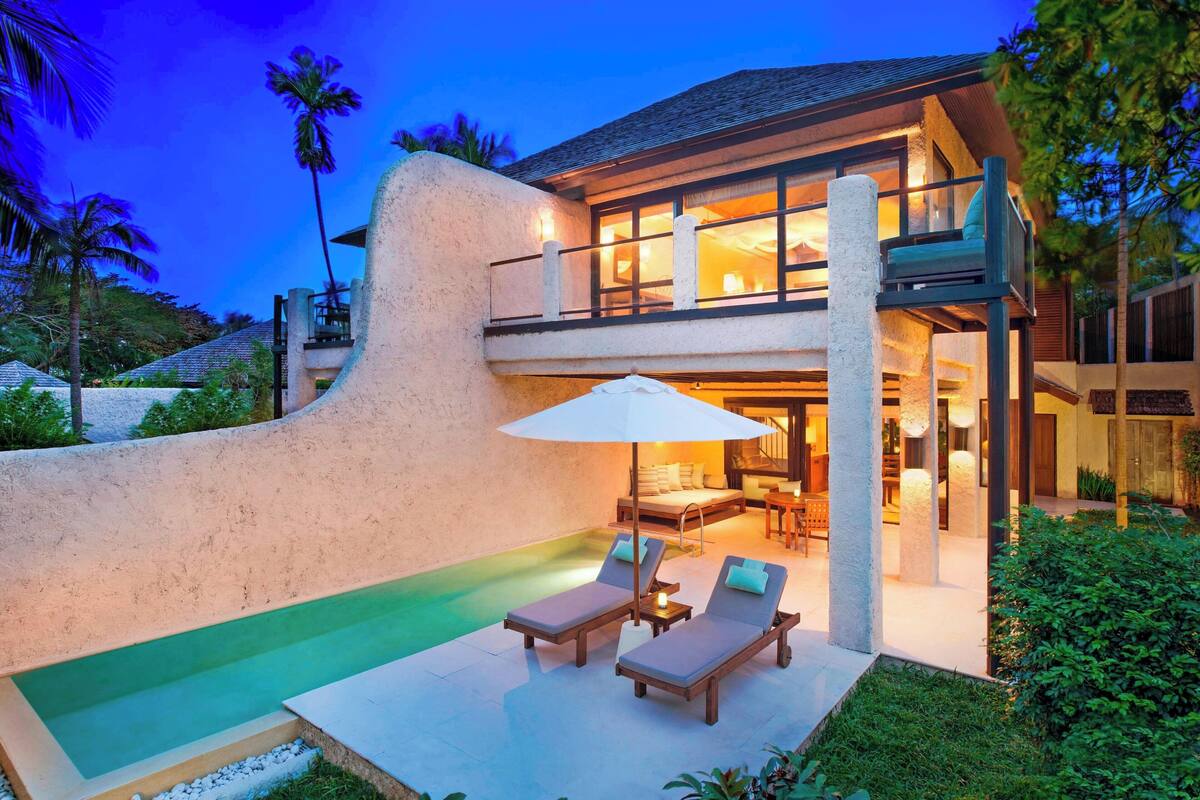 A modern house with a pool, lounge chairs, and a well-lit interior.