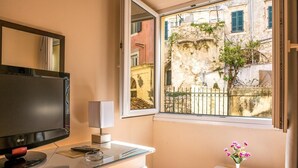 Una habitación con escritorio, televisor, una mesita con un florero y ventana con vistas a edificios.