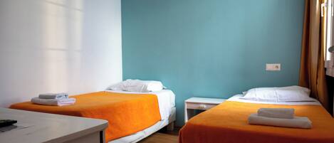 Habitación de hotel con dos camas, cada una con una manta naranja y almohadas blancas, una mesita de noche pequeña y un escritorio con un control remoto.