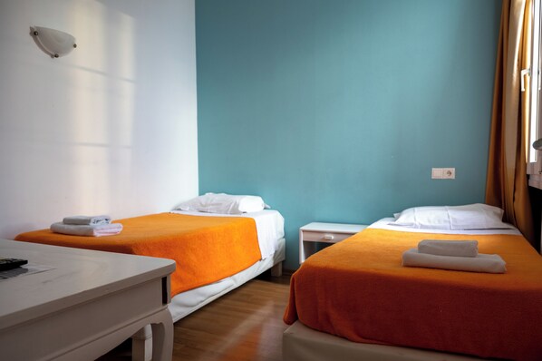 A hotel room with two beds, each with an orange blanket and white pillows, a small bedside table, and a desk with a remote control. - City Marina Hotel (Corfu)