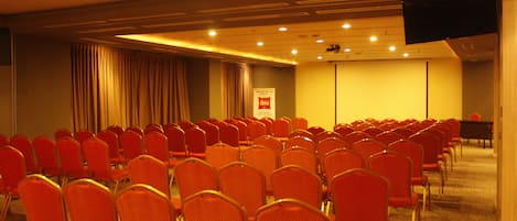 A conference room with rows of red chairs, a projector screen, and a television mounted on the wall.