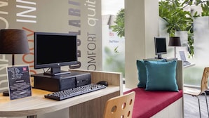 A hotel reception desk with a computer, a keyboard, a desk lamp, a chair, and a window with plants. - ibis Surabaya City Center (Surabaya)