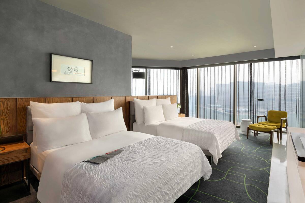 Een hotelkamer met twee bedden, een groot raam met uitzicht over de stad, een lijst aan de muur en een gele stoel.