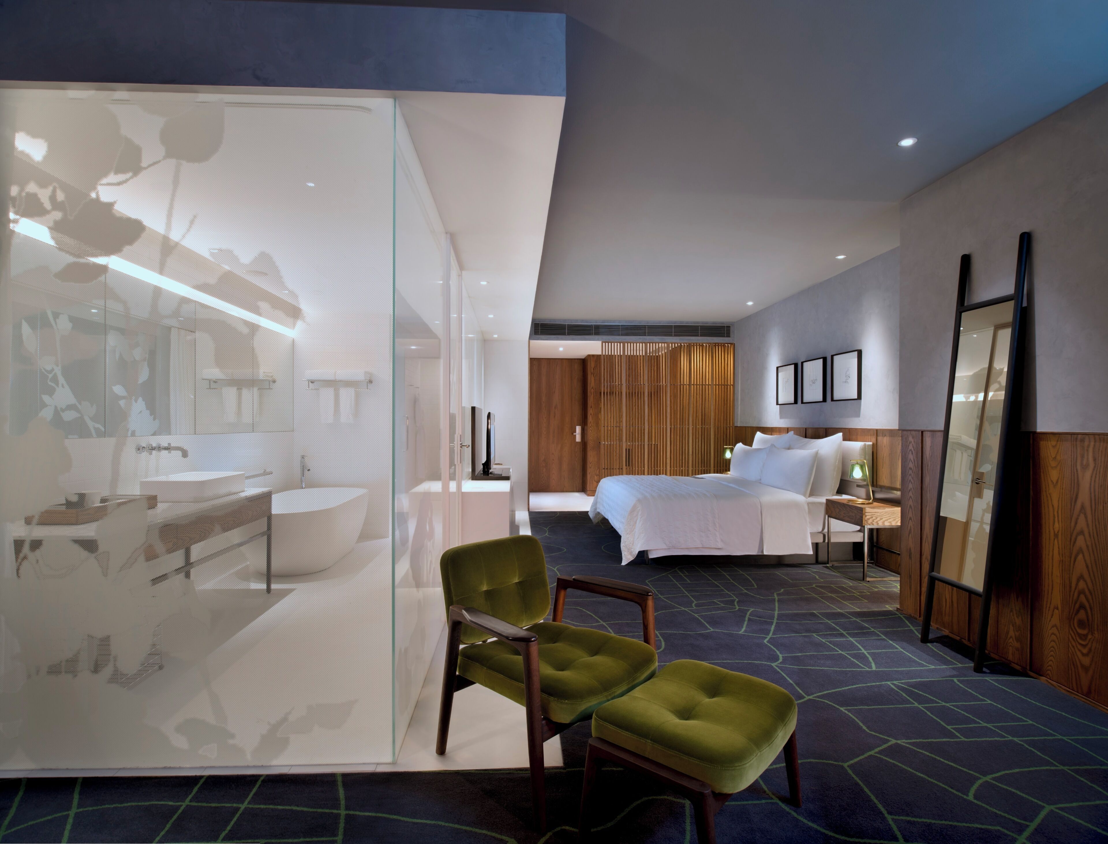 Una habitación de hotel moderna con una cama grande, baño independiente, una silla verde y un espejo amplio.