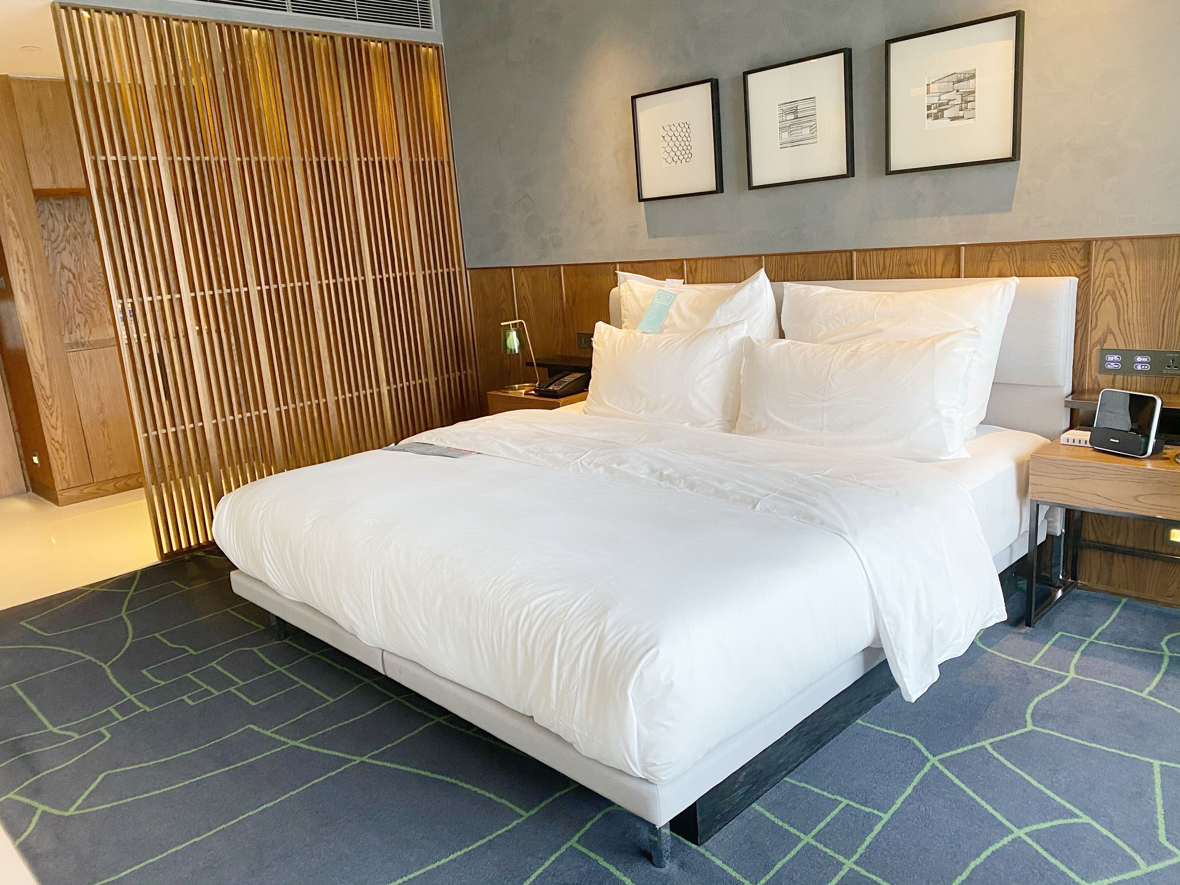 A neatly made bed with white linens, a wooden headboard, a bedside table with a lamp, and a wall-mounted phone.