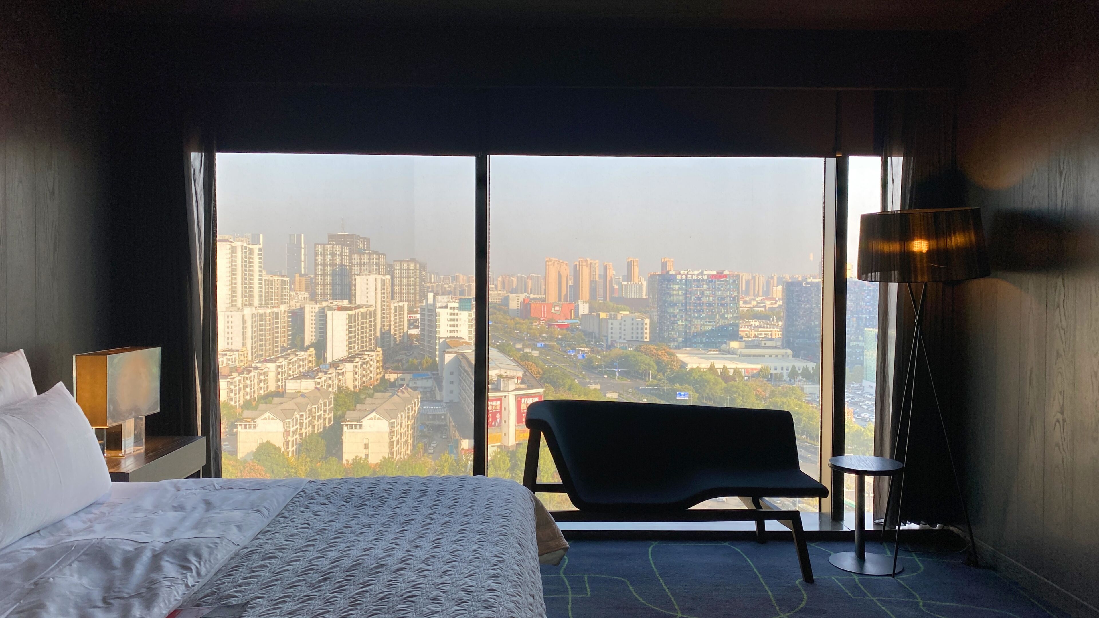 A hotel room with a large bed, a chair, a lamp, and a view of a cityscape through large windows.