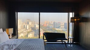 A hotel room with a large bed, a chair, a lamp, and a view of a cityscape through large windows. - Le Méridien Zhengzhou (Zhengzhou)