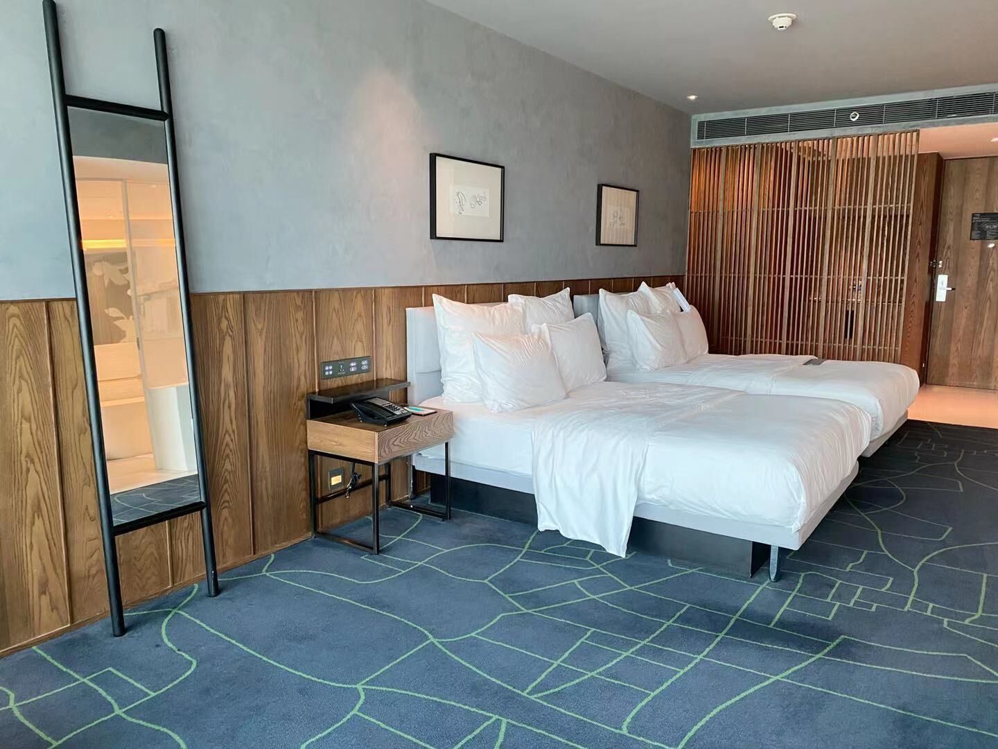 A hotel room with two beds, a bedside table with a phone, a mirror, and framed artwork on the wall.