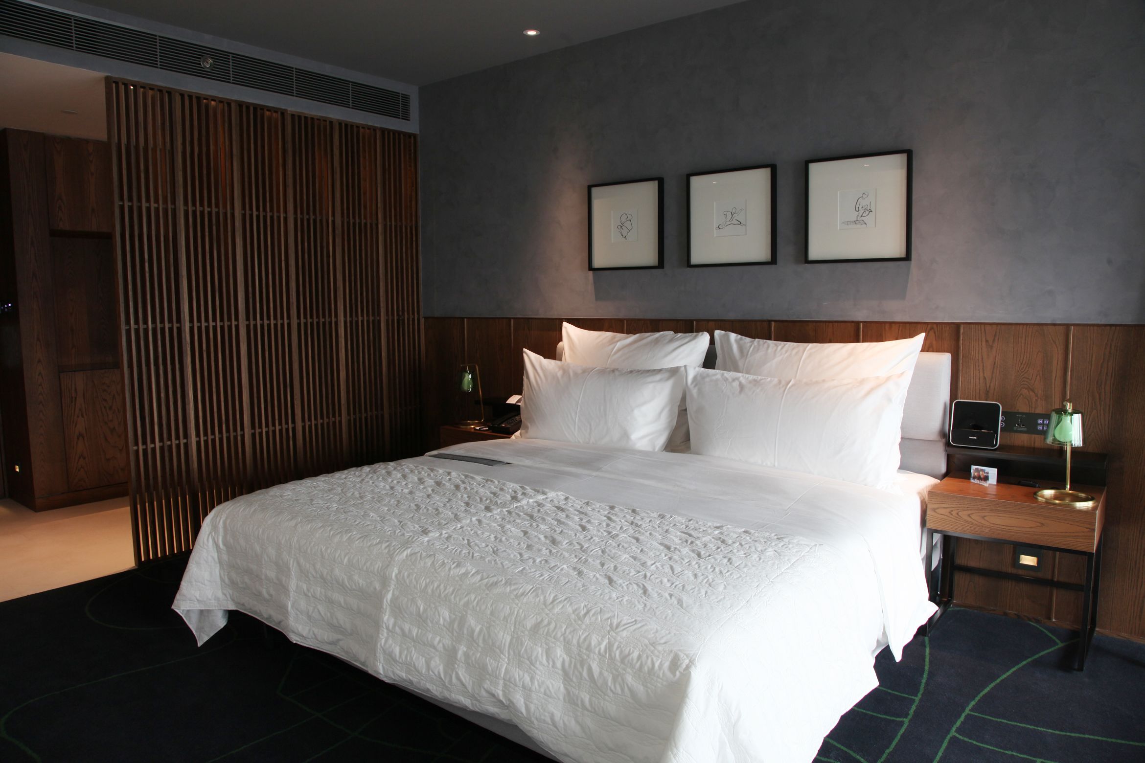 A neatly made bed with white linens, a wooden headboard, and a bedside table with a lamp. There are three framed pictures on the wall above the bed.
