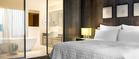 Une chambre d’hôtel avec un grand lit, une douche à parois de verre, une salle de bain avec baignoire et des œuvres d’art encadrées au mur.