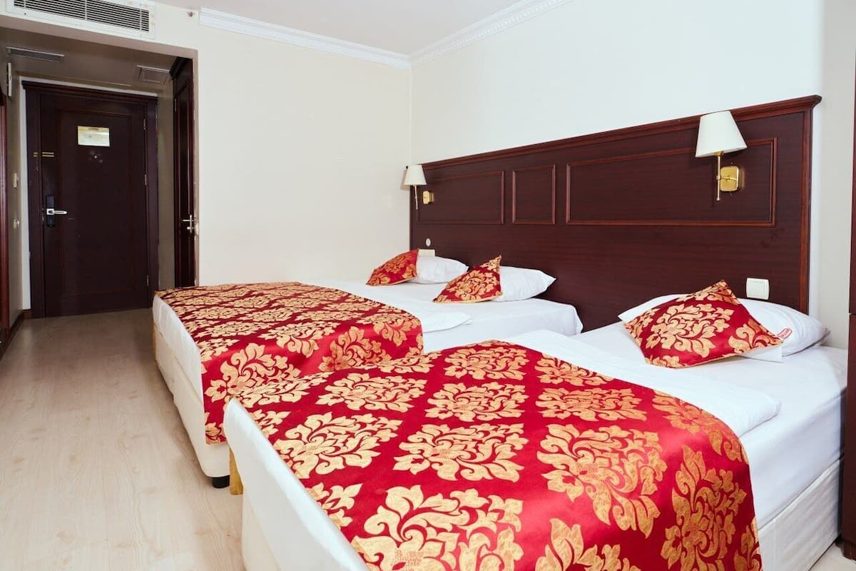 A hotel room with two beds, each with red and gold patterned bedspreads, white pillows, and a wooden headboard.