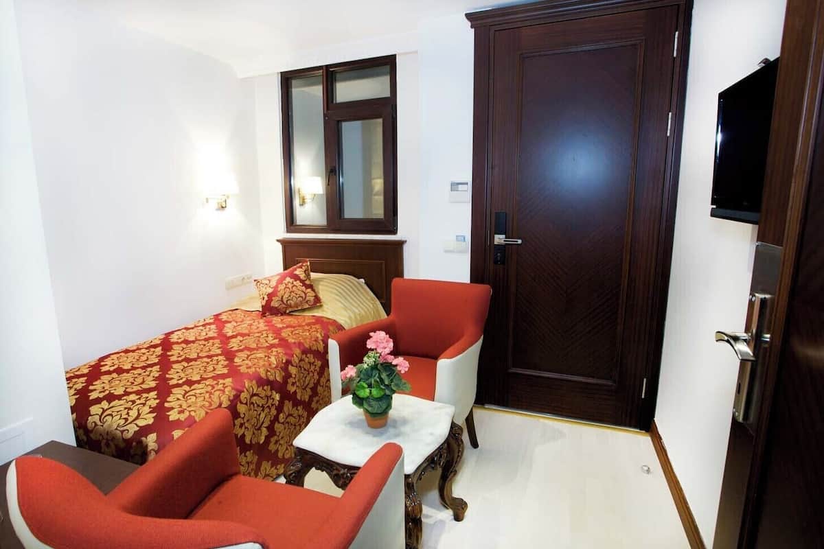A hotel room with a bed, a red armchair, a white table with a vase, a wooden door, and a television.