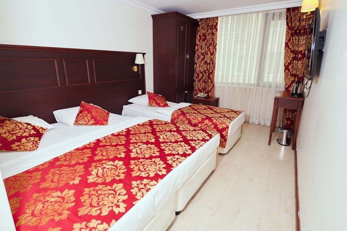 A hotel room with two beds, each with red and gold bedding, a wooden headboard, a small table with a lamp, and a window with curtains.