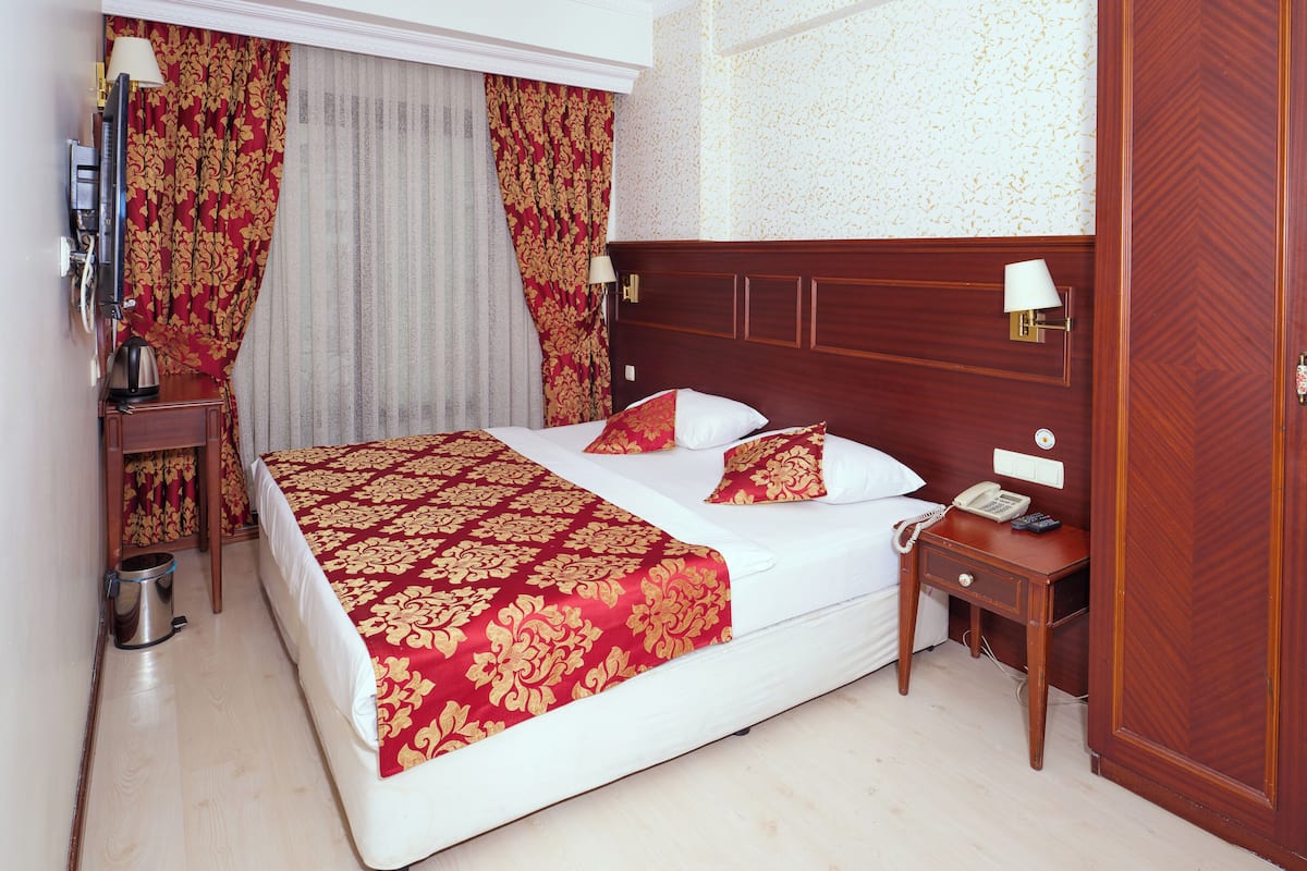 A neatly made bed with a red and gold patterned bedspread, a wooden headboard, a bedside table with a lamp, and a telephone.