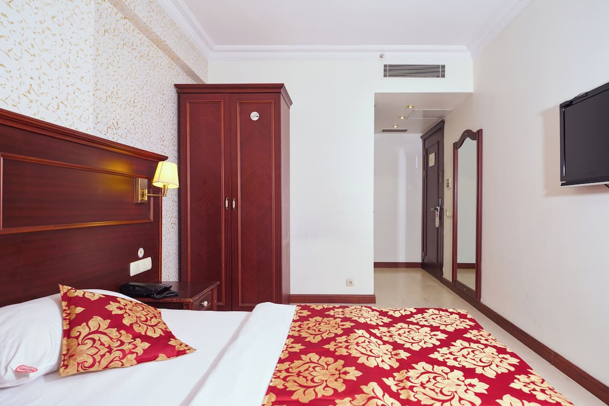 A hotel room with a bed, a wardrobe, a desk with a lamp, and a television on the wall.