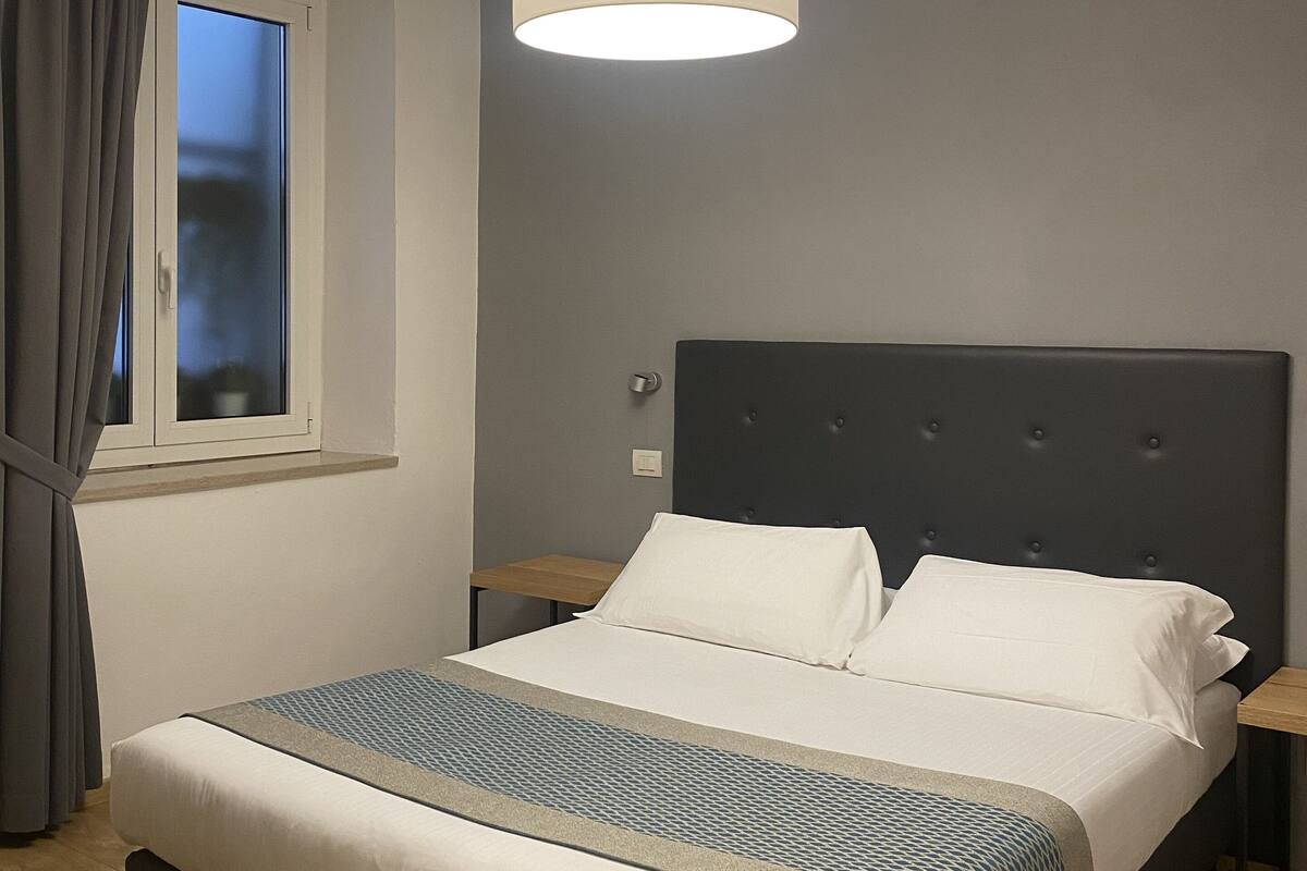 A bedroom with a bed, a bedside table, a window with curtains, and a hanging light fixture.
