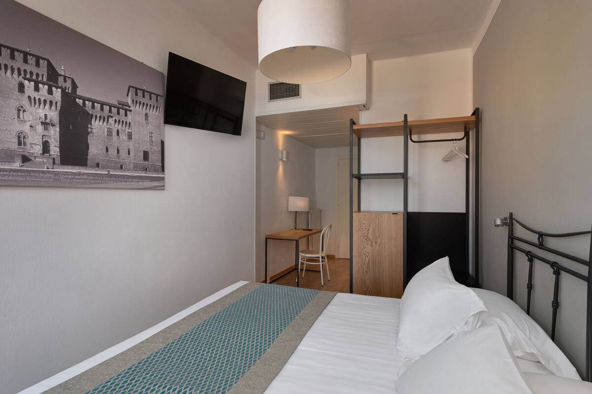 A hotel room with a bed, a desk, a chair, a coat rack, and a television mounted on the wall.