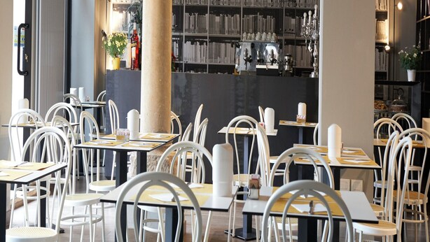 Un café moderne situé dans une bibliothèque, avec des chaises et des tables blanches, des étagères et une grande lampe de lecture.