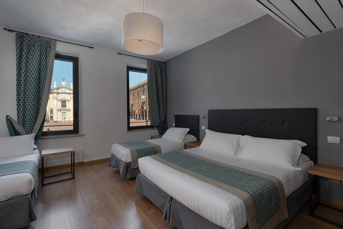A hotel room with two beds, a window with a view of a building, a hanging light fixture, and a wooden floor.