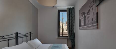 A bedroom with a bed, a window with a view of buildings, a hanging light fixture, and a framed picture on the wall.