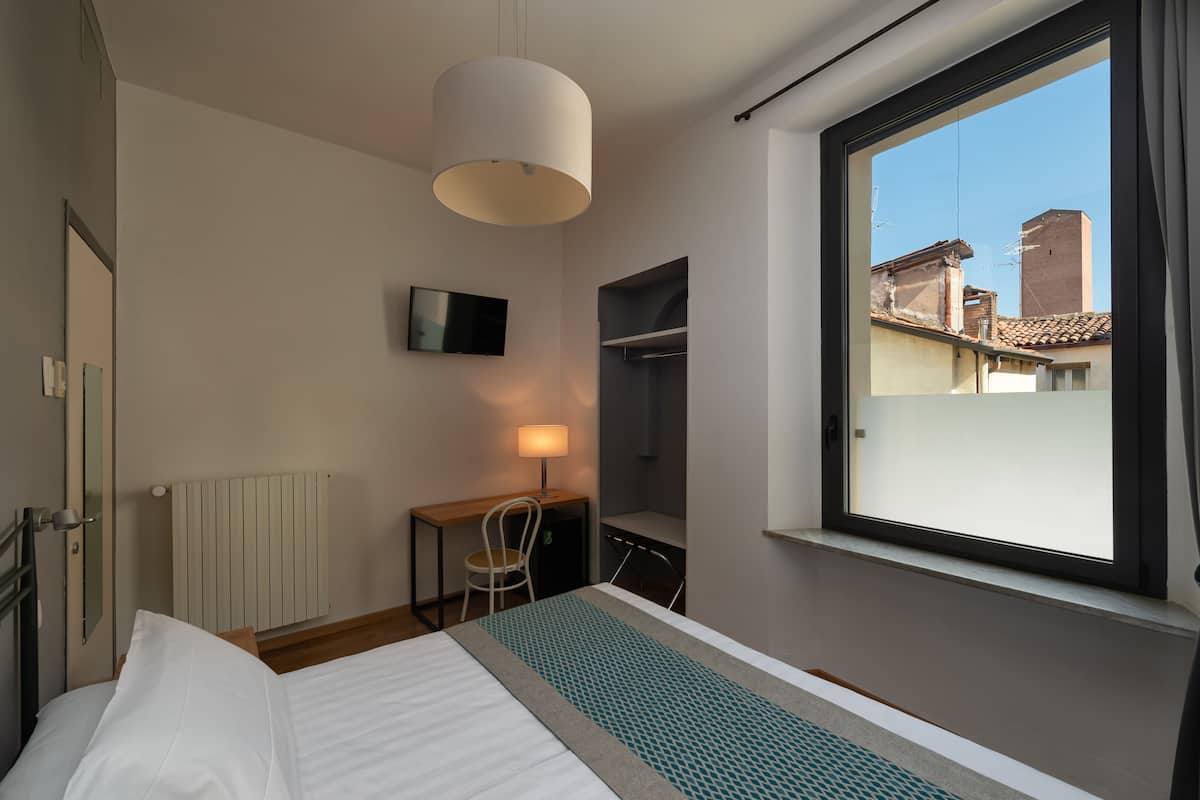 A hotel room with a bed, a desk with a chair, a TV, a window with a view of buildings, and a hanging light fixture.