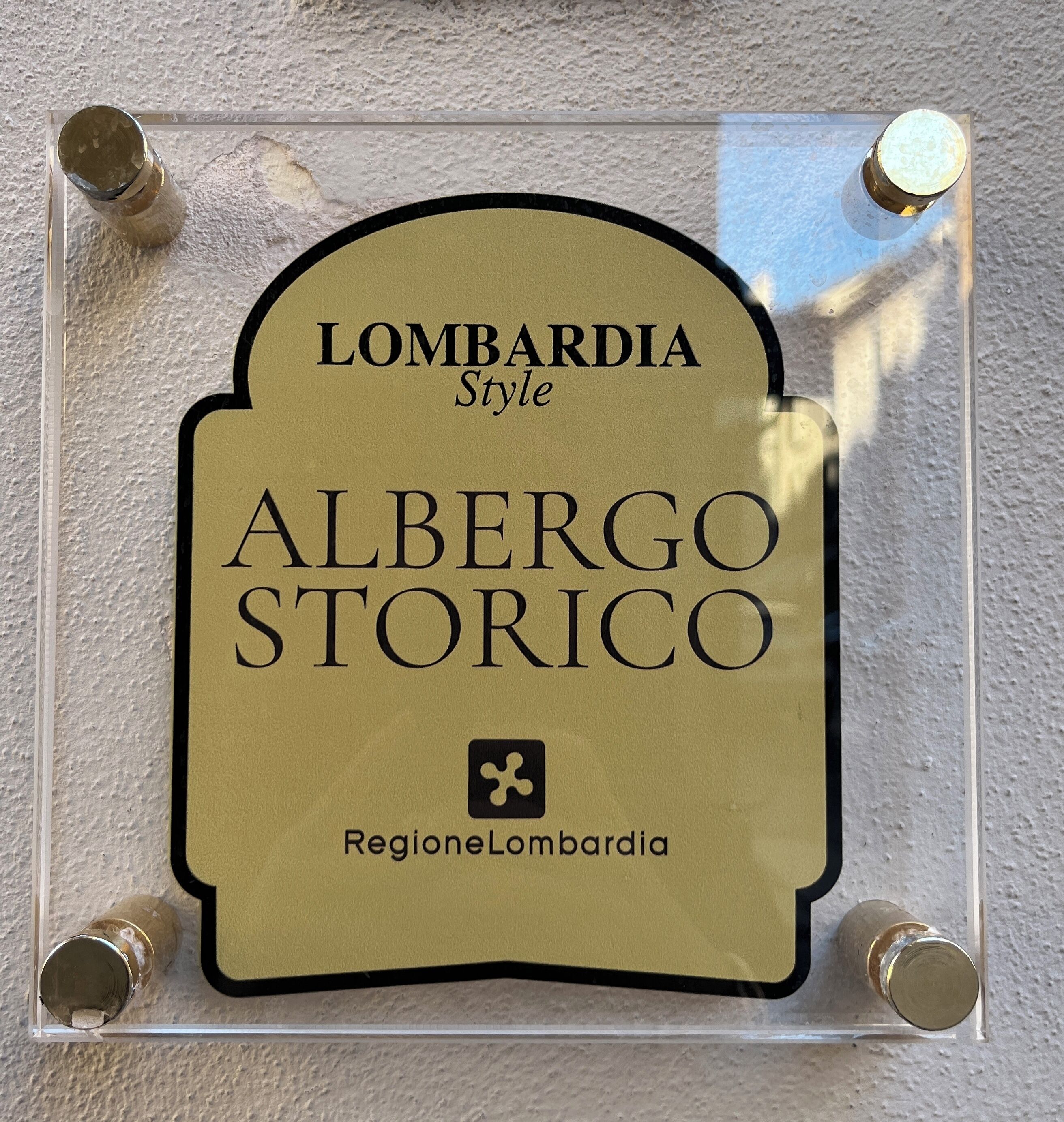 Letrero con el texto 'Lombardia Style' y 'Alberto Storico' sobre un fondo beige, fijado a la pared con cuatro tornillos.