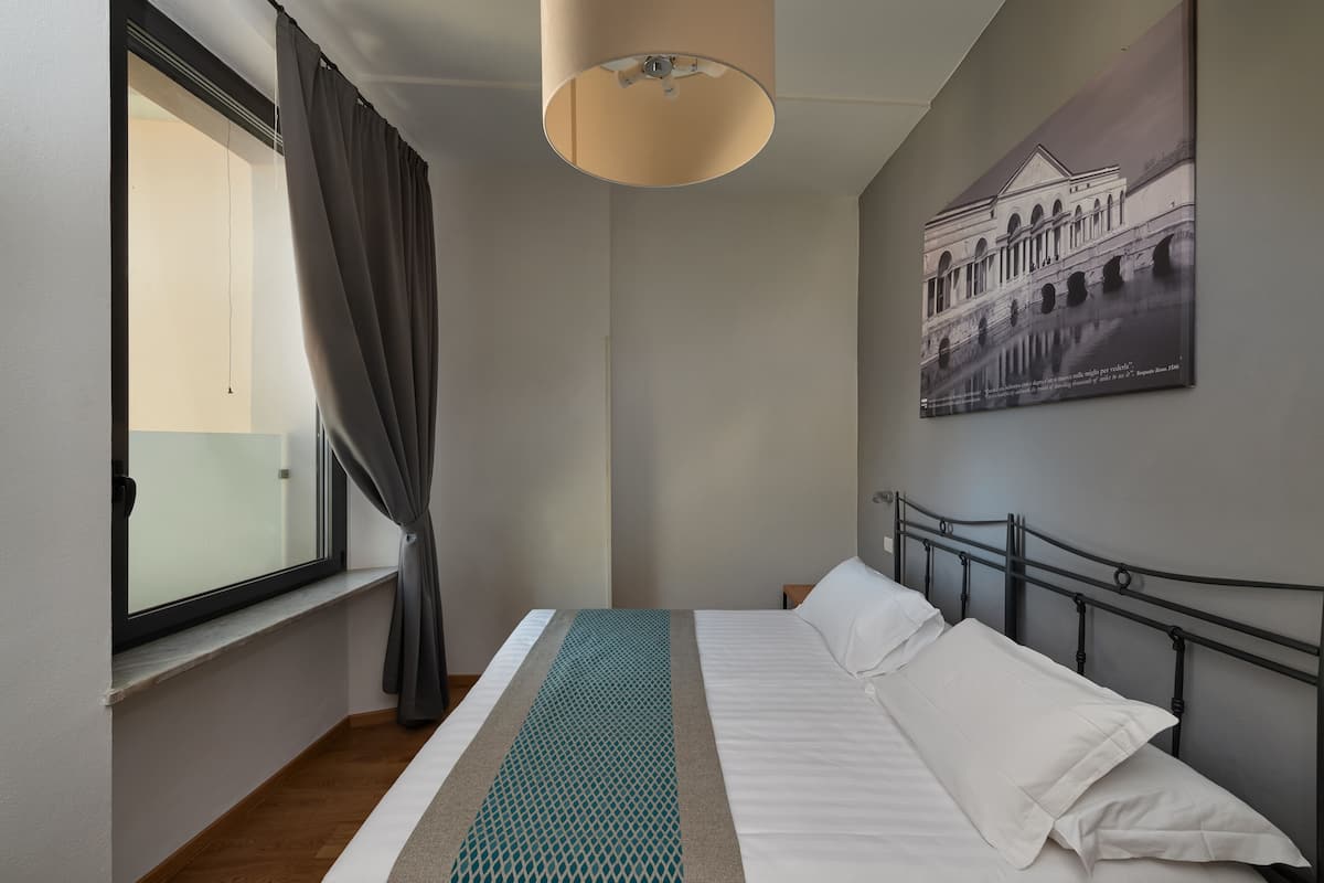 A neatly made bed with white linens, a metal headboard, and a teal patterned throw. A large window with curtains is visible, and a wall-mounted lamp hangs above the bed.