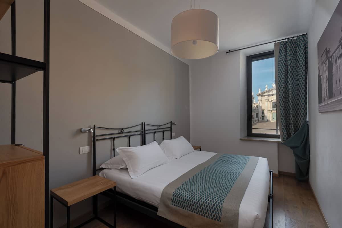 A bedroom with a bed, a bedside table, a hanging light fixture, a window with a view of buildings, and a curtain.
