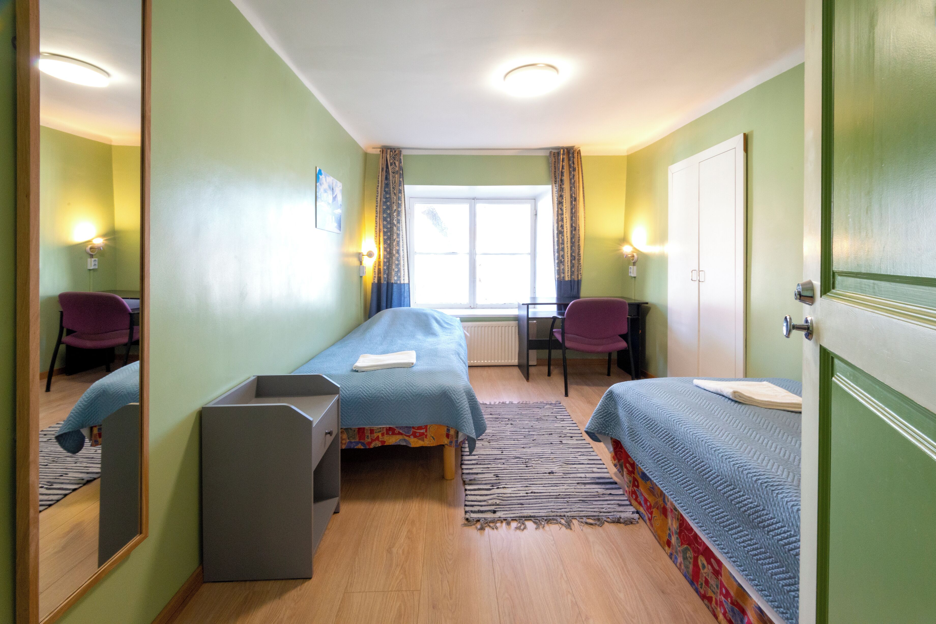 Une chambre avec deux lits simples, un bureau, une chaise, un miroir et une fenêtre avec des rideaux.