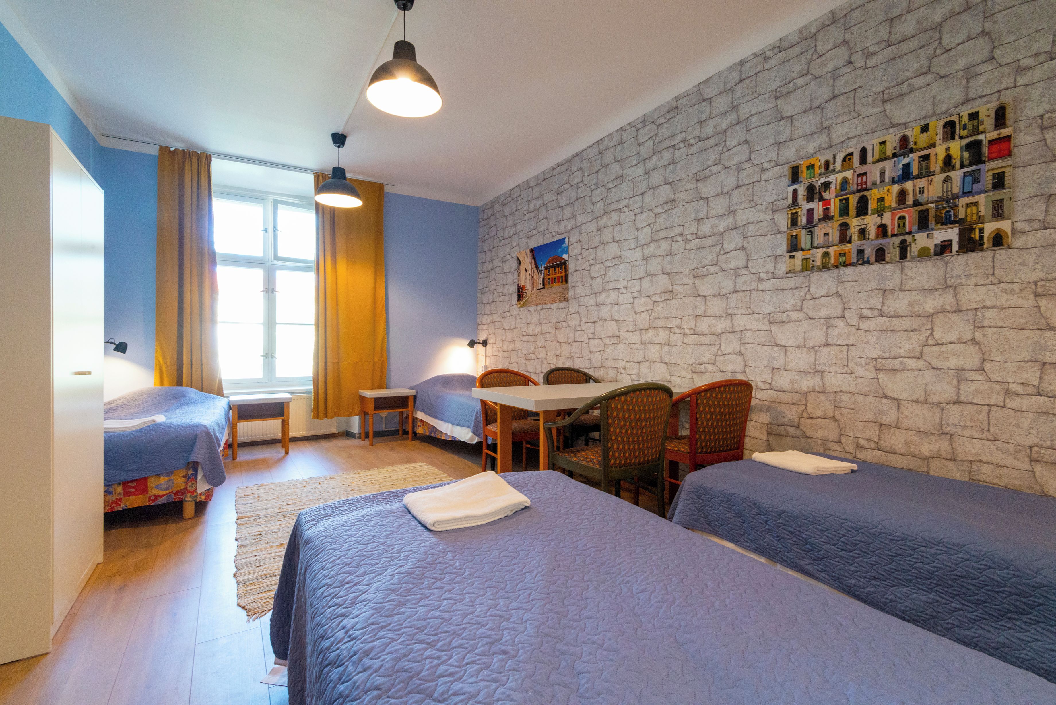 Una habitación con dos camas, una mesa, sillas, un ventanal con cortinas y una pared de piedra con un mural colorido.