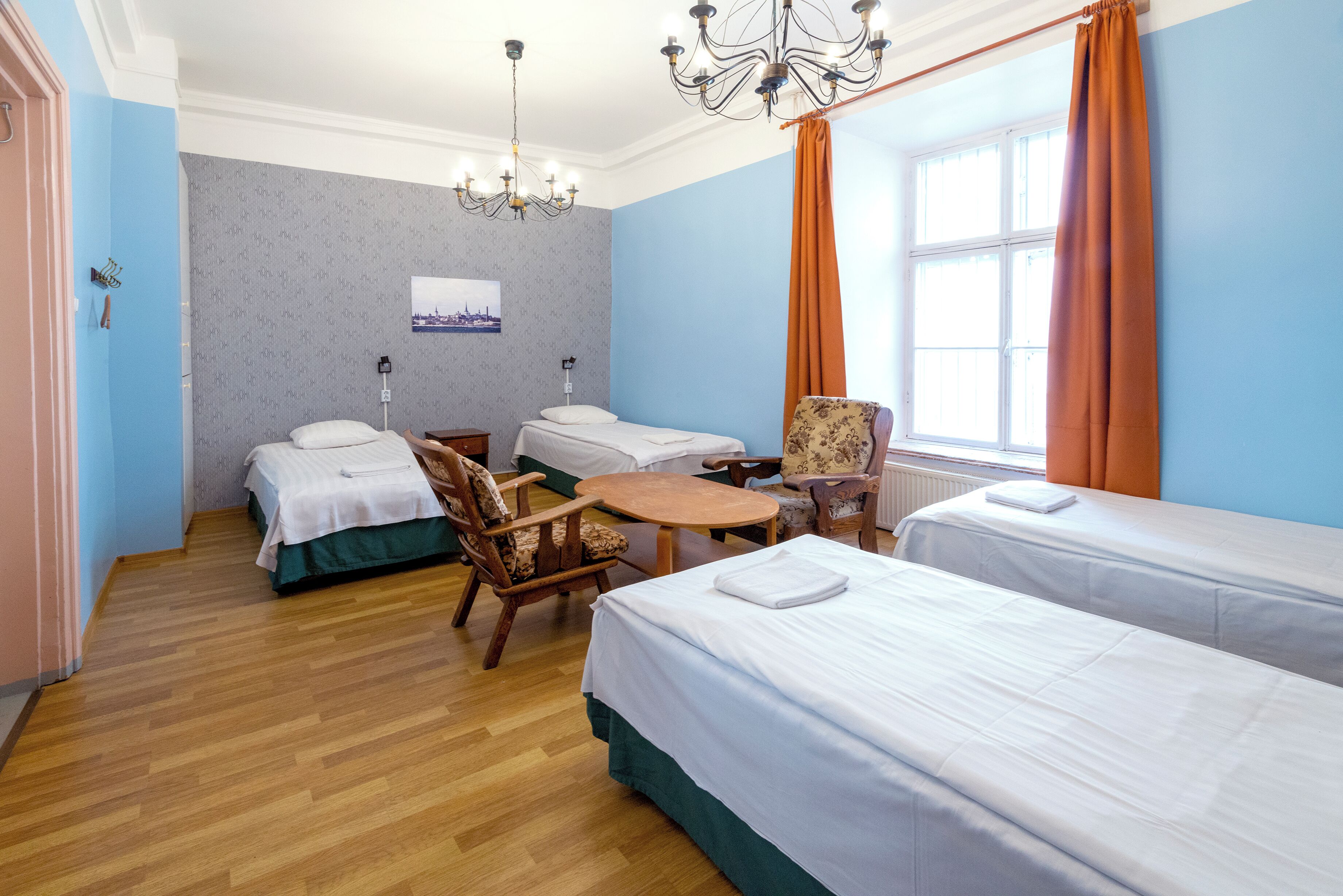 A hotel room with two beds, a wooden chair, a small table, a chandelier, and a window with curtains.