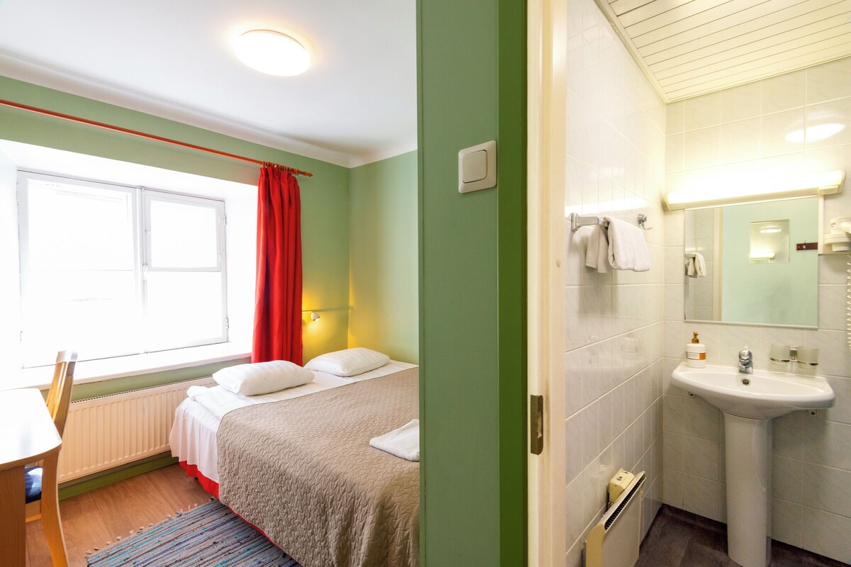 A hotel room with a bed, a desk, a window with red curtains, and a bathroom with a sink and mirror.