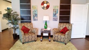 A living room with two patterned armchairs, a small table with a lamp, and a large potted plant.