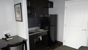 A compact kitchen with a refrigerator, microwave, and sink. There is a chair and a small table with a phone on it. A door is visible to the right.