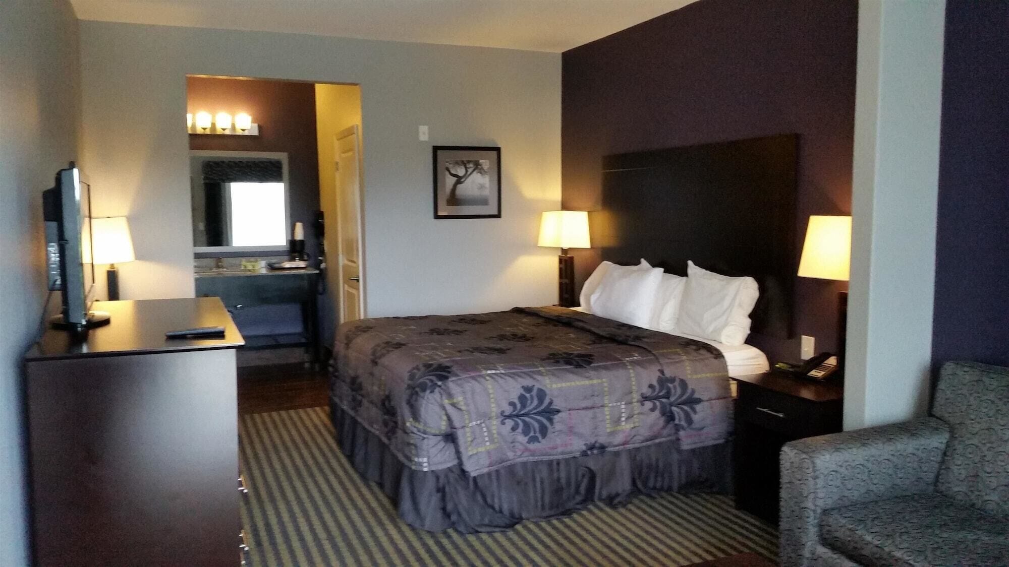 A hotel room with a bed, a desk with a lamp, a chair, a TV, and a bathroom visible in the background.