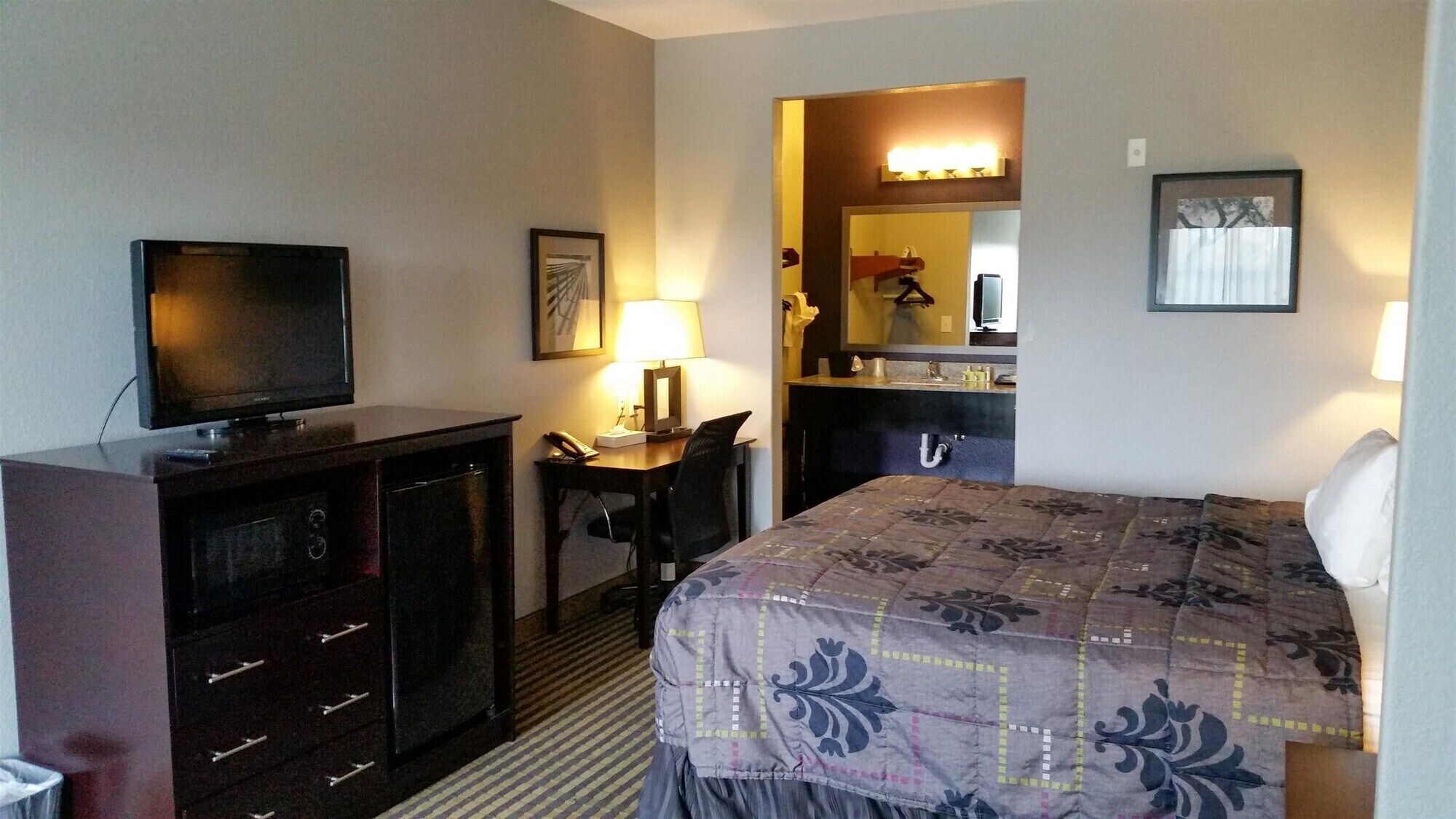 A hotel room with a bed, a desk with a chair, a television, a dresser, a mirror, and a bathroom with a sink and mirror.